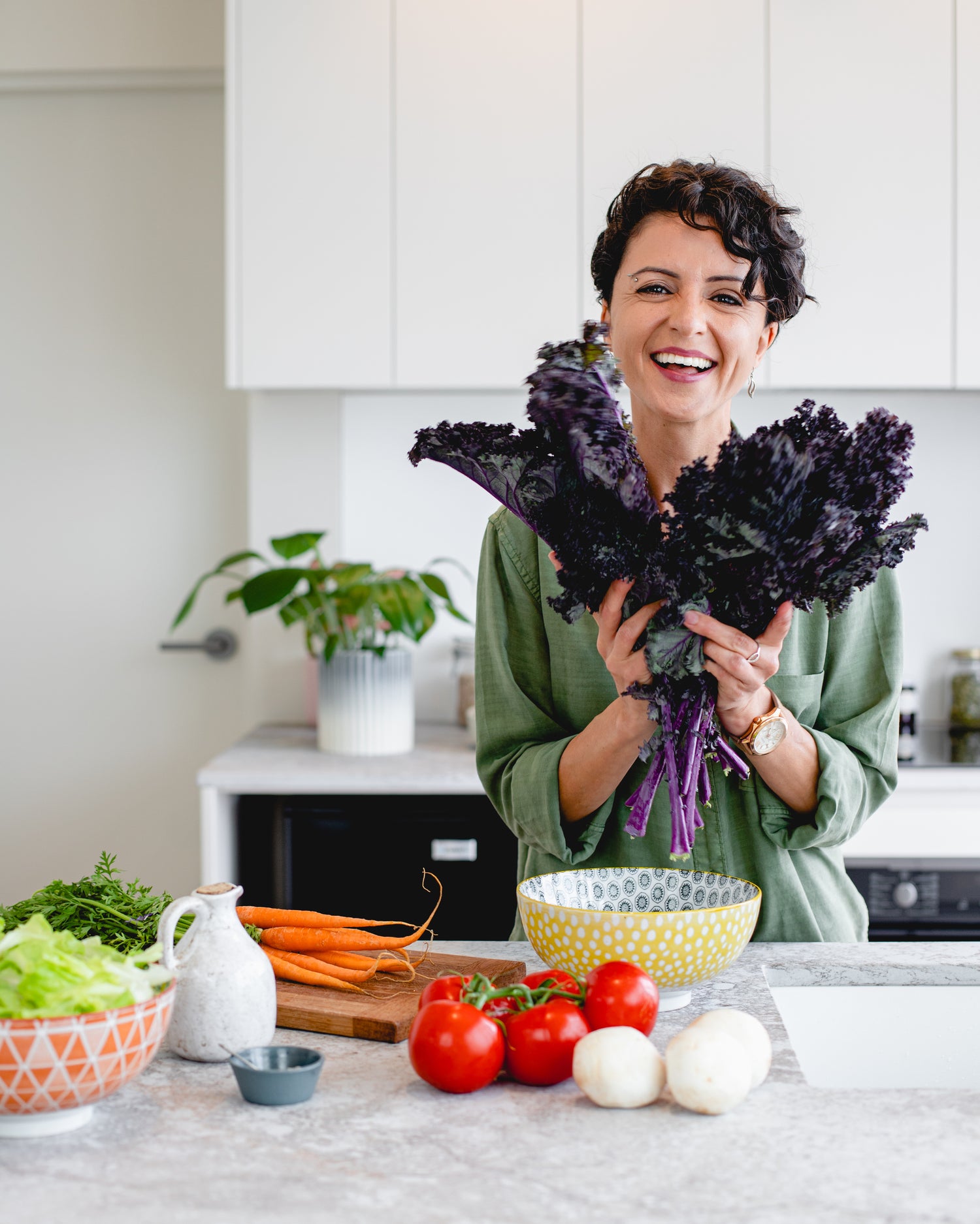 White woman holding lettuce in her hand in a kitchen with a table full of healthy and nutritious foods, including tomatoes and carrots.