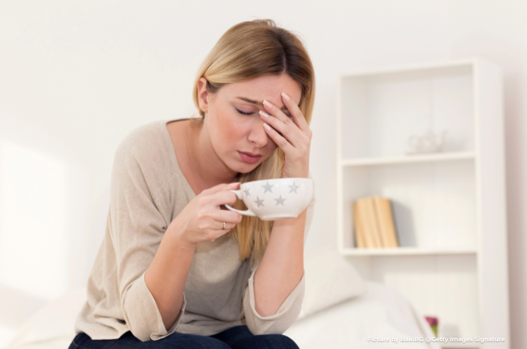 A tired-looking woman in her 40s experiencing symptoms of perimenopause and PMDD, such as a headache or fatigue, while sitting on a sofa holding a cup of tea.